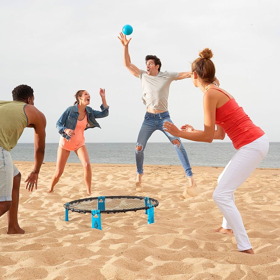 Four people play Franklin Spyderball by Franklin on a sandy beach near the ocean. One player leaps to hit the blue ball while others cheer, ready to catch it. The sky is partly cloudy.