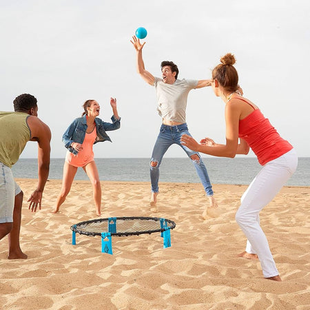 Four people play Franklin Spyderball by Franklin on a sandy beach near the ocean. One player leaps to hit the blue ball while others cheer, ready to catch it. The sky is partly cloudy.