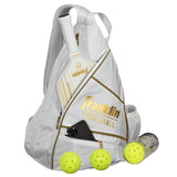 The Franklin Pickleball Sling Bag by Franklin comes in White/Gold and holds a paddle, white towel, smartphone, and water bottle. Three yellow pickleballs sit in front of this stylish gear bag.