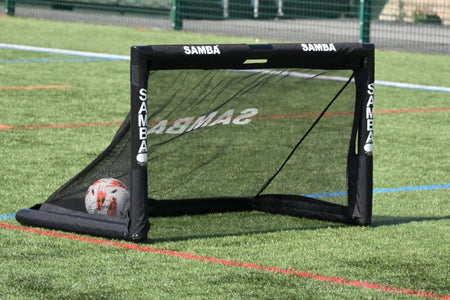 A Samba Sports 4ft x 2.5ft Folding 3v3 Goal with a black frame is set on artificial turf. A soccer ball sits inside the net, while blue and red field lines mark the pitch—perfect for quick 3v3 games or training sessions.