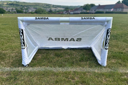 A Samba Sports 4ft x 2.5ft Folding 3v3 Goal with a white frame stands on a grassy field, with houses, trees, and rolling hills in the background beneath a partly cloudy sky.