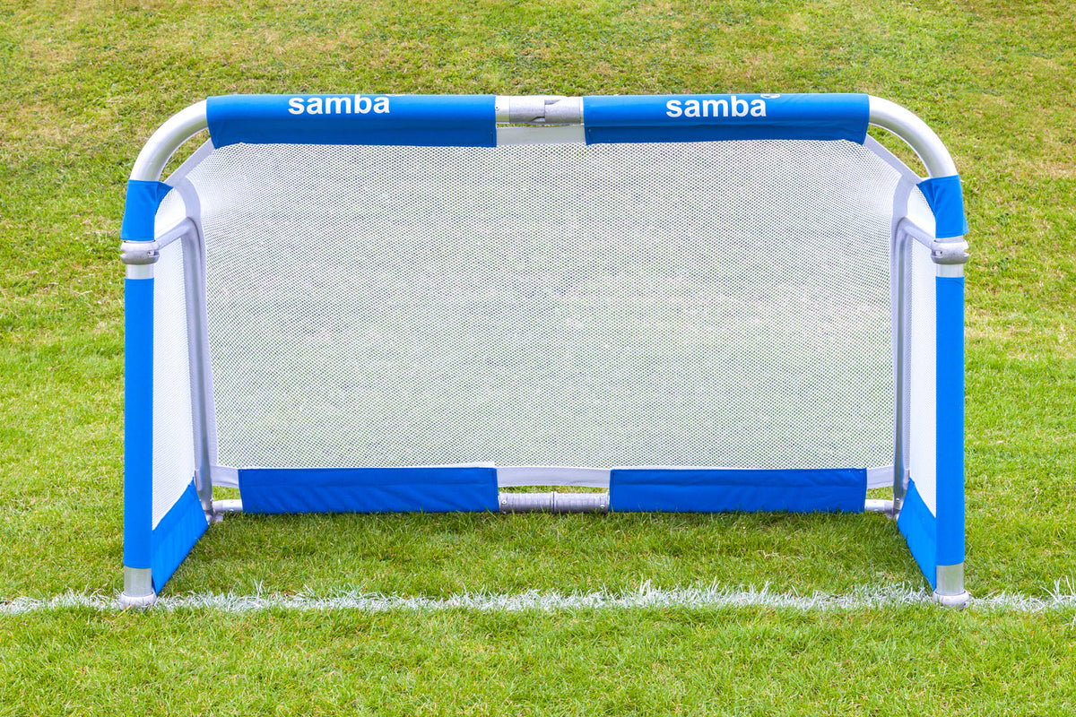 A Samba Sports Aluminium Folding Football Goal (5ft x 3ft) with blue and white accents stands on a grassy field, featuring the word “Samba” printed twice on the top padding. Its foldable aluminium design ensures easy transport and quick setup for games wherever you need.
