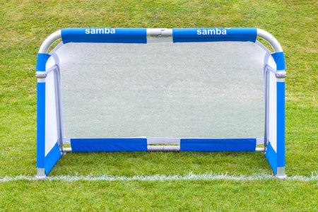 A Samba Sports Aluminium Folding Football Goal (5ft x 3ft) with blue and white accents stands on a grassy field, featuring the word “Samba” printed twice on the top padding. Its foldable aluminium design ensures easy transport and quick setup for games wherever you need.