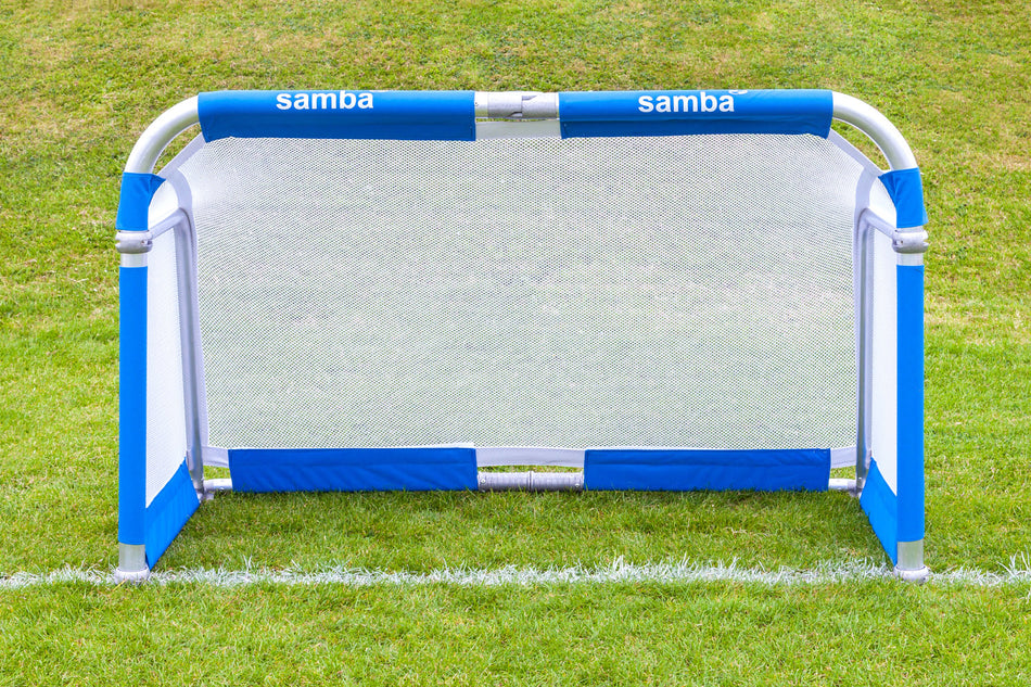 A compact and portable Samba Aluminium Folding Football Goal (5ft x 3ft) by Samba Sports, featuring a white net and blue frame, is set up on green grass near a white line, with “Samba Aluminium Folding Football Goal” displayed on the top padding. This lightweight goal is easy to move and ideal for quick games anywhere.