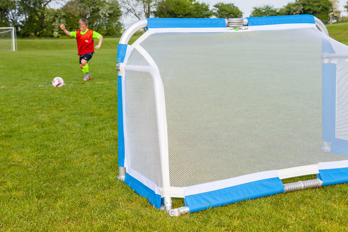 A child in a red shirt runs toward a Samba Aluminium Folding Football Goal (5ft x 3ft) by Samba Sports on a grassy field, preparing to kick a soccer ball. Trees and another football goal are visible in the background.