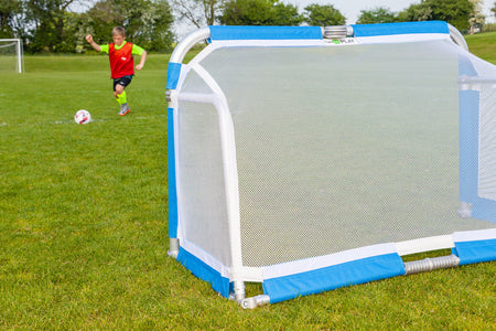 A child in a red shirt runs toward a Samba Aluminium Folding Football Goal (5ft x 3ft) by Samba Sports on a grassy field, preparing to kick a soccer ball. Trees and another football goal are visible in the background.