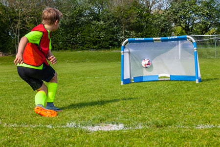 A young boy in sportswear kicks a soccer ball towards a Samba Sports Samba 3v3 4ft x 2.5ft Aluminium Folding Football Goal on a grassy field with trees behind.