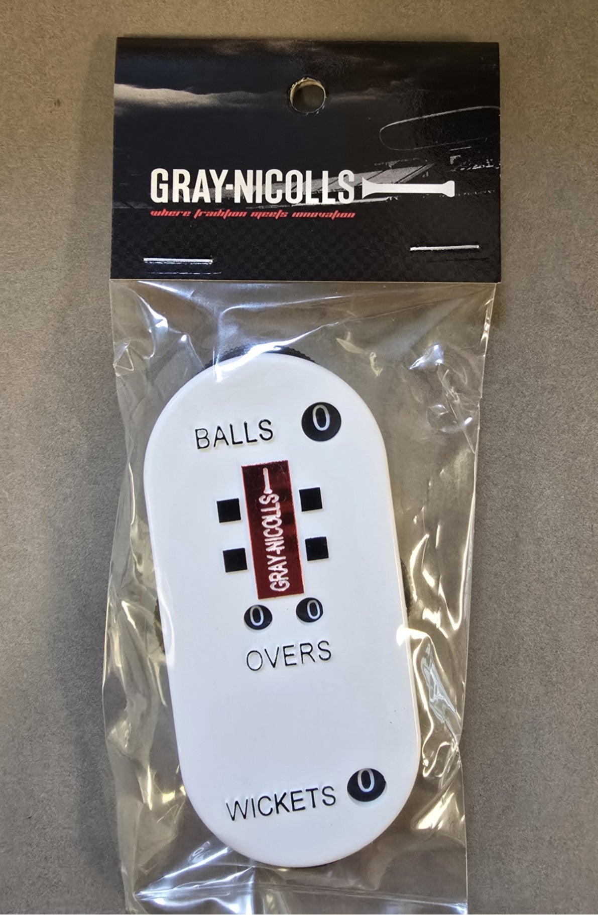 The Gray Nicolls Umpires Counter by Gray Nicolls is packaged, features dials for tracking balls, overs, and wickets (all set to zero), and rests on a grey surface.
