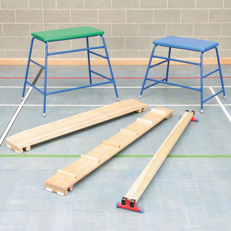 Two Niels Larsen Agility Set benches, one green and one blue, stand behind two timber balance beams on a gym floor with colored lines, displaying quality Niels Larsen gymnastic equipment against a brick wall.