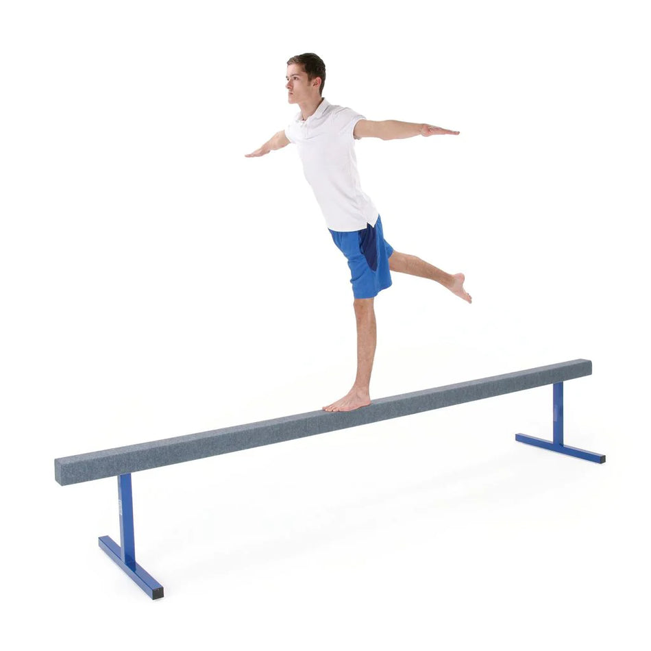 A person balances barefoot on one leg on a Niels Larsen Training Beam (61cm high), wearing a white shirt and blue shorts, with arms extended and the other leg raised behind. The timber beam with steel legs is shown against a plain white background.