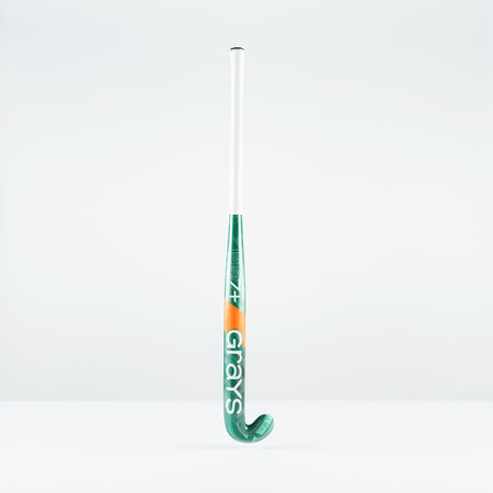 A green and white Grays PB 7+ Junior Hockey Stick 2026 with orange accents stands upright against a plain white background, displaying the brand name and offering intermediate power plus supreme control.