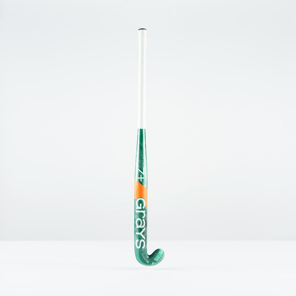 A green and white Grays PB 7+ Junior Hockey Stick 2026 with orange accents stands upright against a plain white background, displaying the brand name and offering intermediate power plus supreme control.