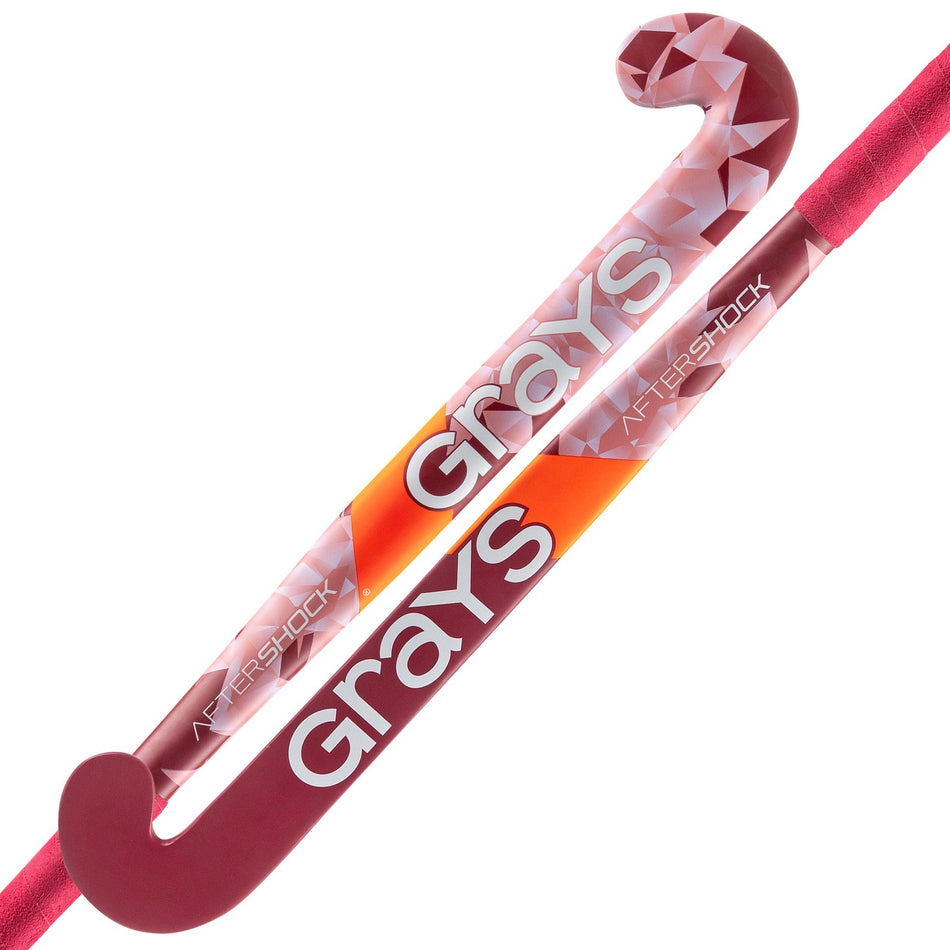 Two pink/red Grays Aftershock Wooden Hockey Sticks are displayed side by side, each with a classic bow shape, geometric patterns, and the large white GRAYS branding along the shafts.