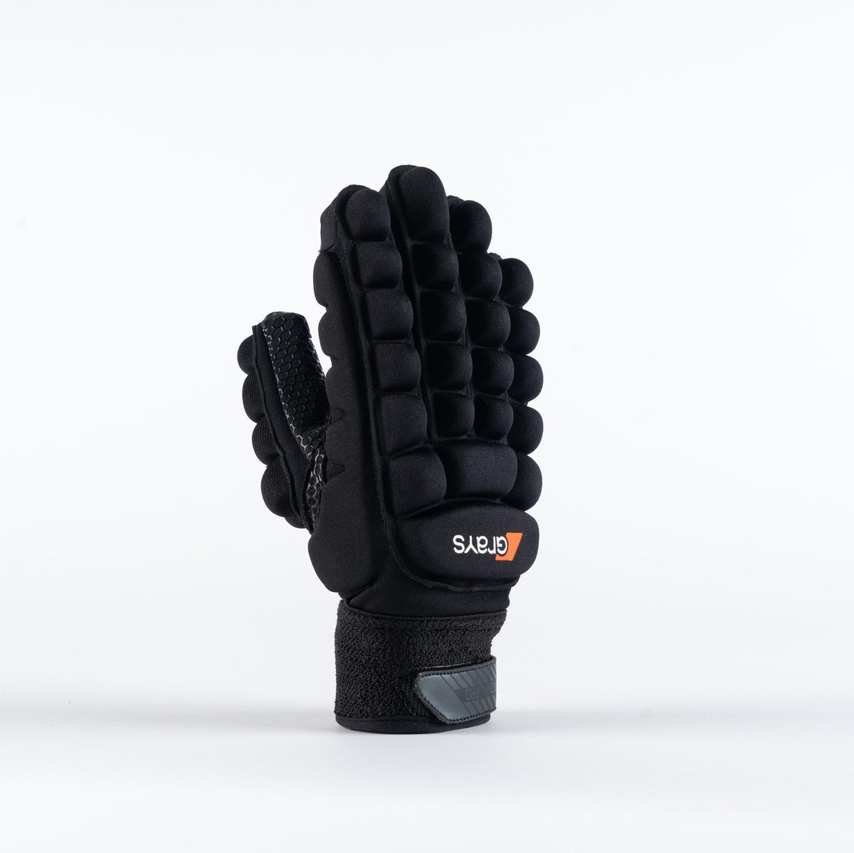 The Grays International Pro Hockey Glove by Grays, in black, is shown standing upright on a plain white background. It features a lycra palm, thick segmented cushioning on the back, Velcro wrist strap, and an orange and white logo.