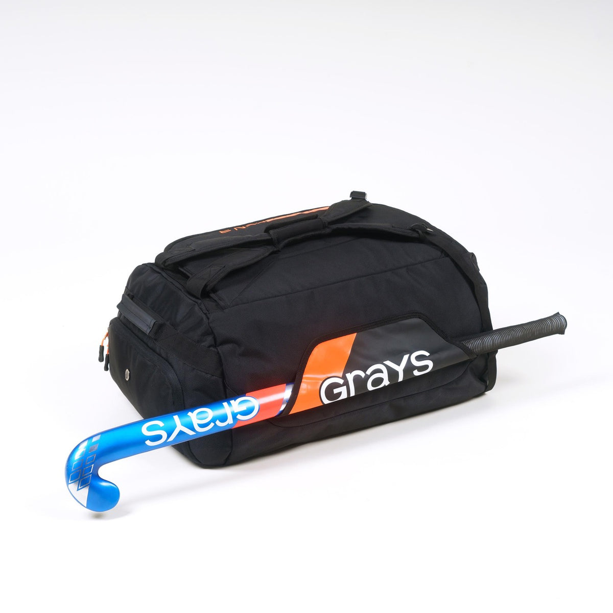 The Grays Omega Gen 3 Holdall, a black bag with orange accents and partially visible Grays branding, holds a blue and orange Grays field hockey stick and features a dedicated shoe pocket, all set against a white background.