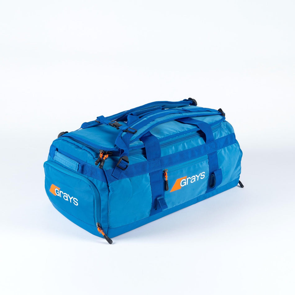 The Grays Omega Gen 4 Holdall is a spacious blue hockey gear bag with the Grays logo in white and orange, multiple zippered compartments, handles, adjustable shoulder straps, and a stick-ready design—ideal for pro players.