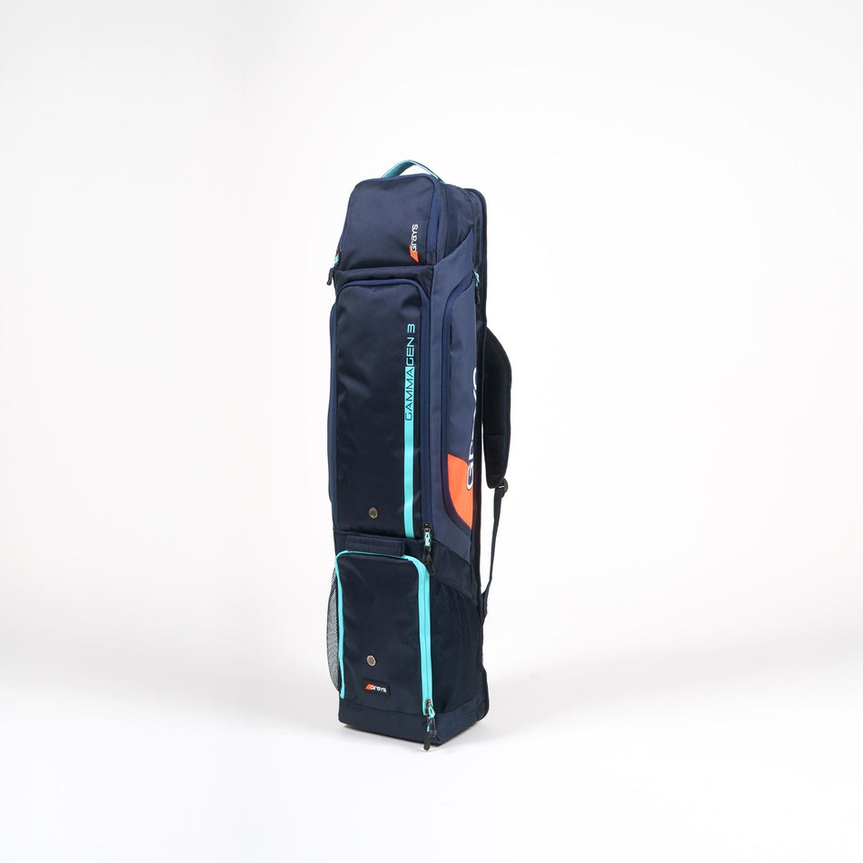 The Grays Gamma Gen3 Hockey Kitbag by Grays, in Navy/Aqua, features water-resistant material, light blue zippers, orange accents, multiple compartments, and a dedicated sticks section. It stands upright on a plain white background.