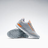 The Grays Raid 2.0 Hockey Shoes by Grays are shown in grey on a white background—one upright, one tilted to reveal the textured sole's aggressive grip system and striking orange accents for high performance on the field.