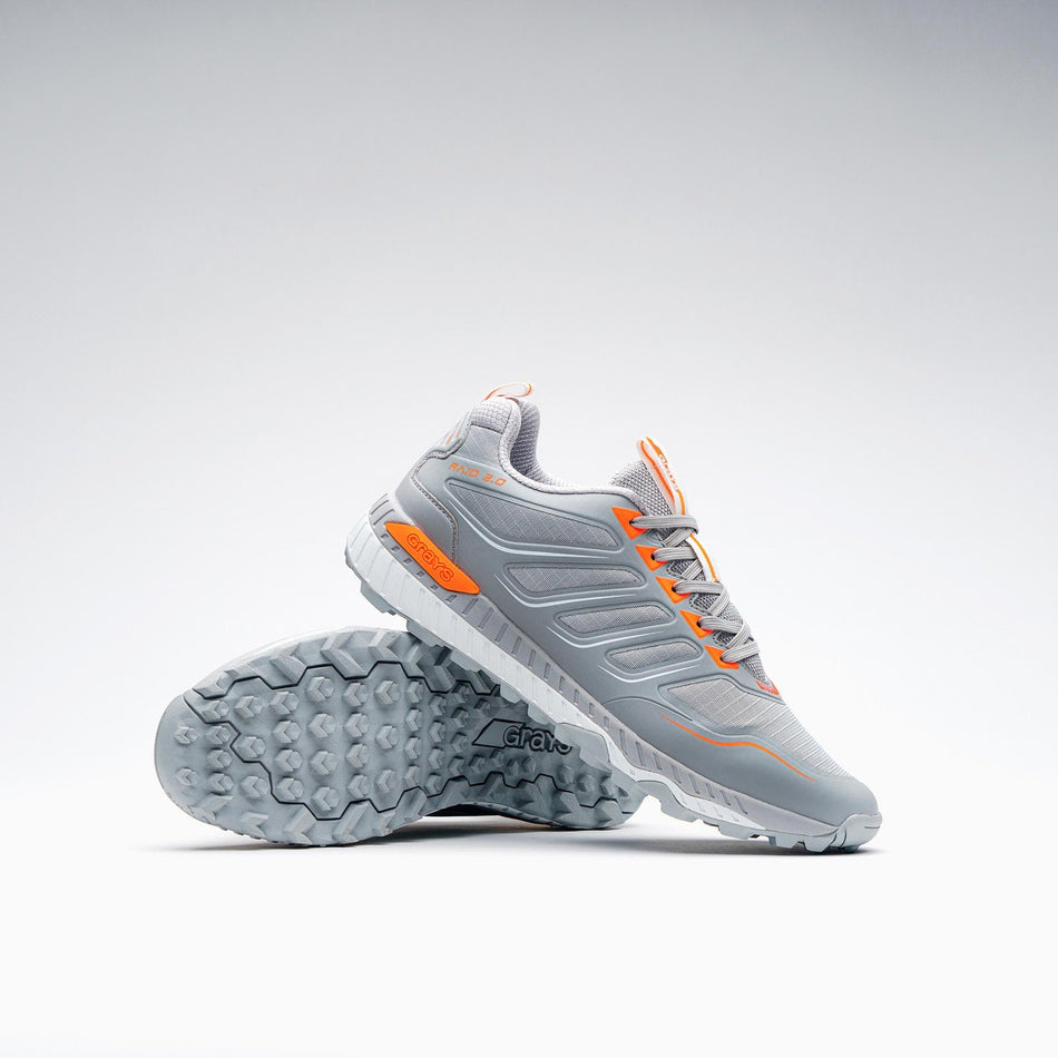 The Grays Raid 2.0 Hockey Shoes by Grays are shown in grey on a white background—one upright, one tilted to reveal the textured sole's aggressive grip system and striking orange accents for high performance on the field.