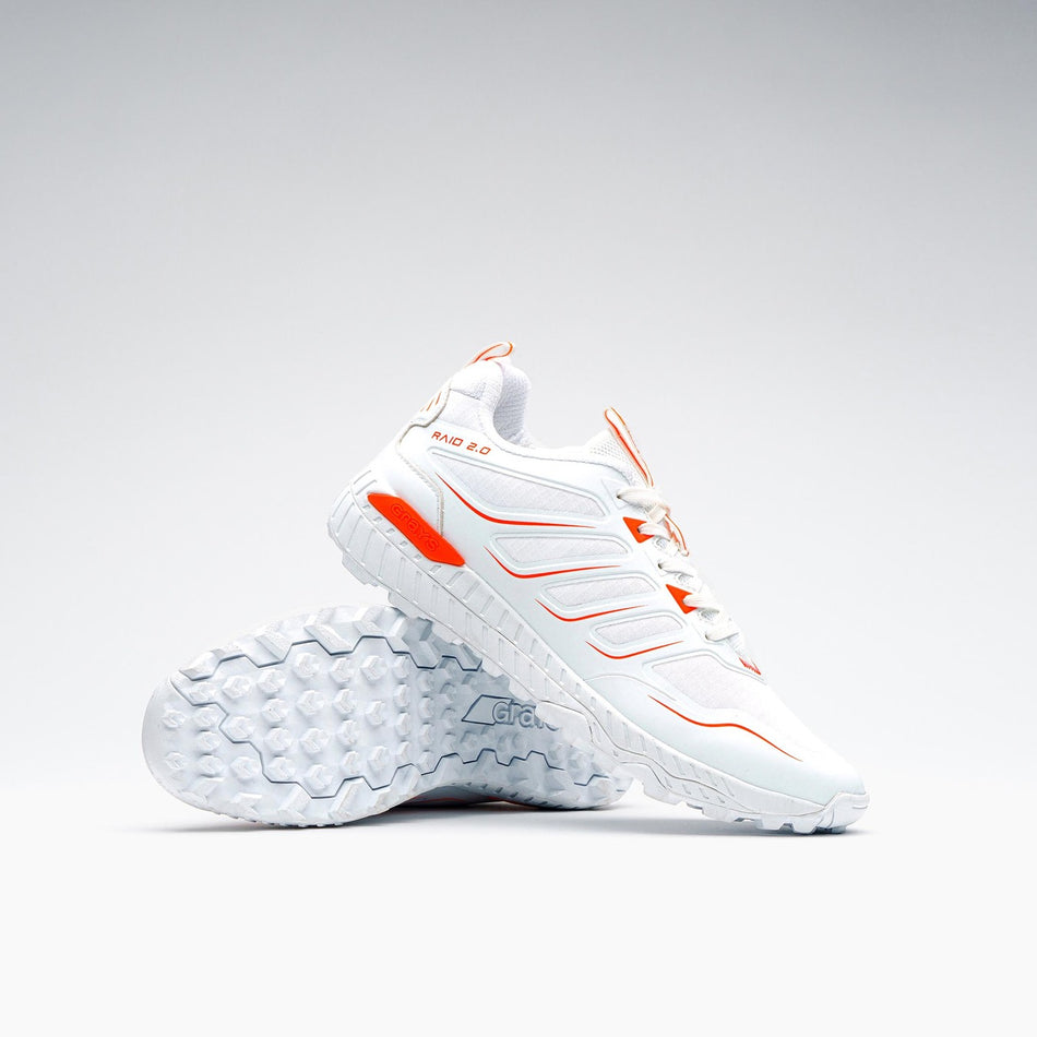 A pair of Grays Raid 2.0 Hockey Shoes by Grays in white are shown: one stands upright, the other leans to display its textured sole and aggressive grip—all set against a plain, light background.