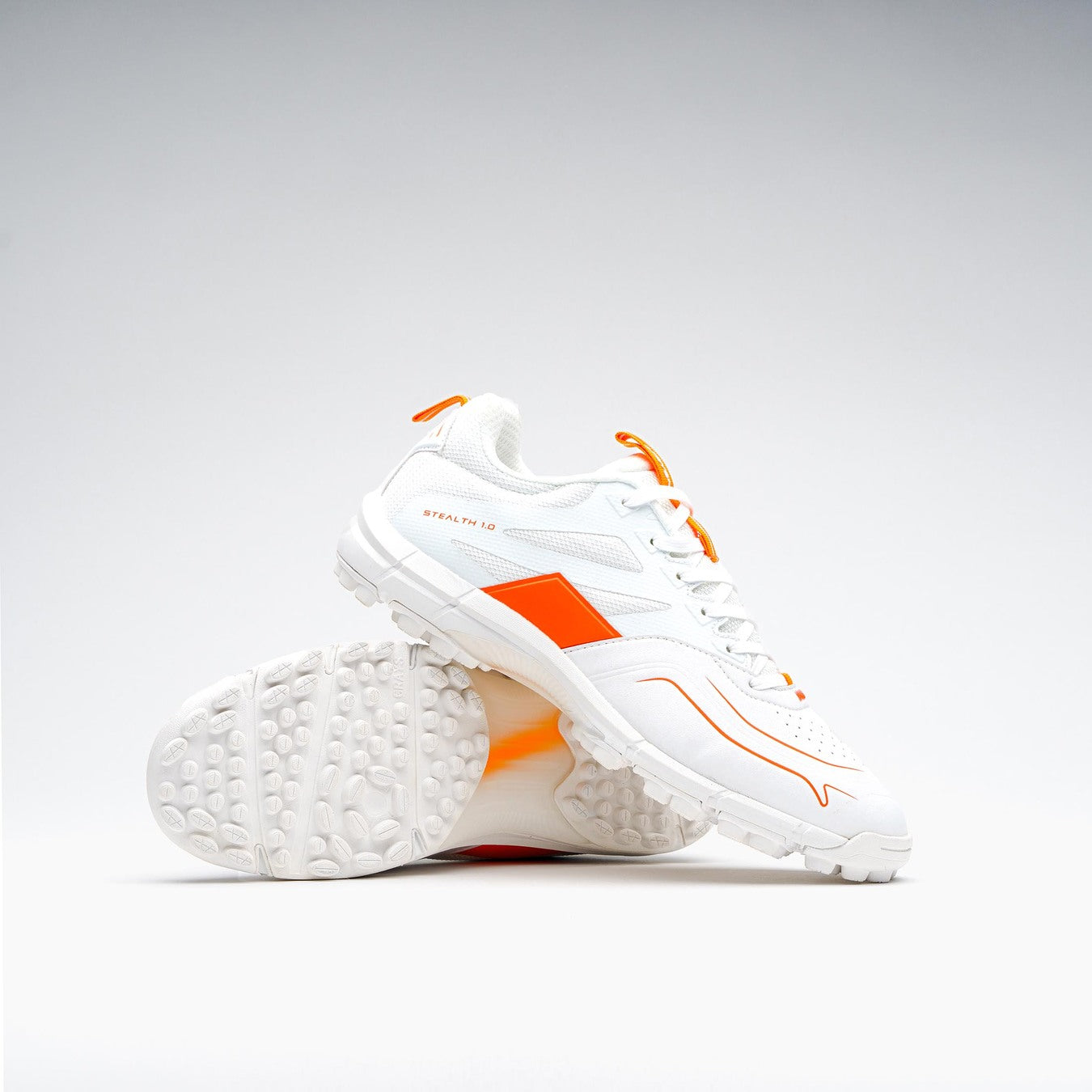 The Grays Stealth 1.0 Hockey Shoes in white, by Grays, are shown on a plain white background—one upright and one on its side to highlight the textured high-performance outsole and orange detailing.