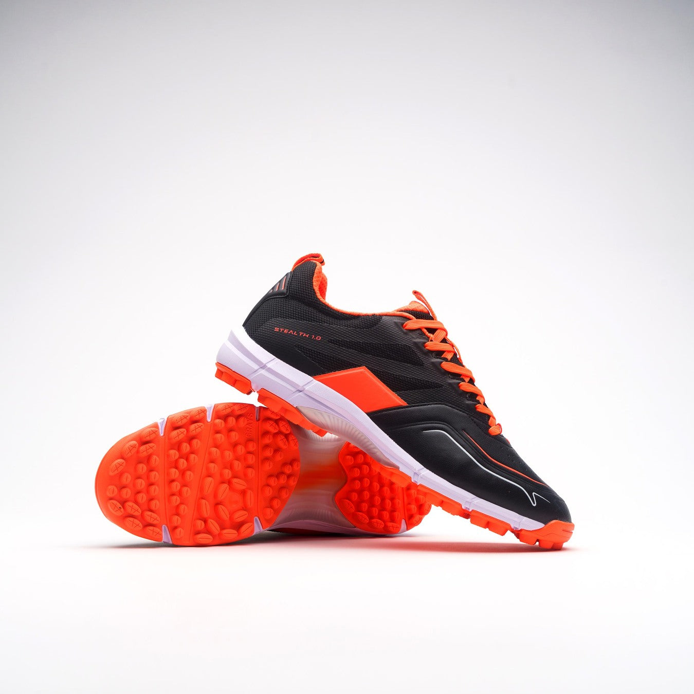 A pair of Grays Stealth 1.0 Hockey Shoes by Grays in Black/Orange, featuring bright orange soles and laces with a high-performance rubber outsole; one shoe upright, the other on its side, shown on a white background.