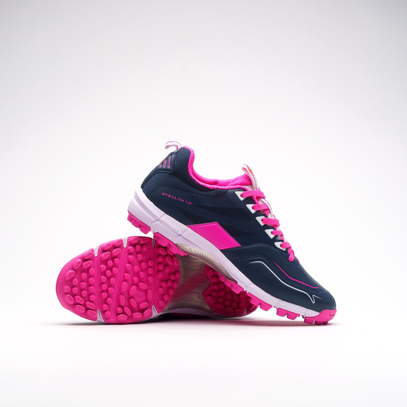 The Grays Stealth 1.0 Hockey Shoes by Grays feature navy uppers, bright pink accents, white soles, and pink textured outsoles. One shoe stands while the other leans to reveal their high-grip performance rubber sole.