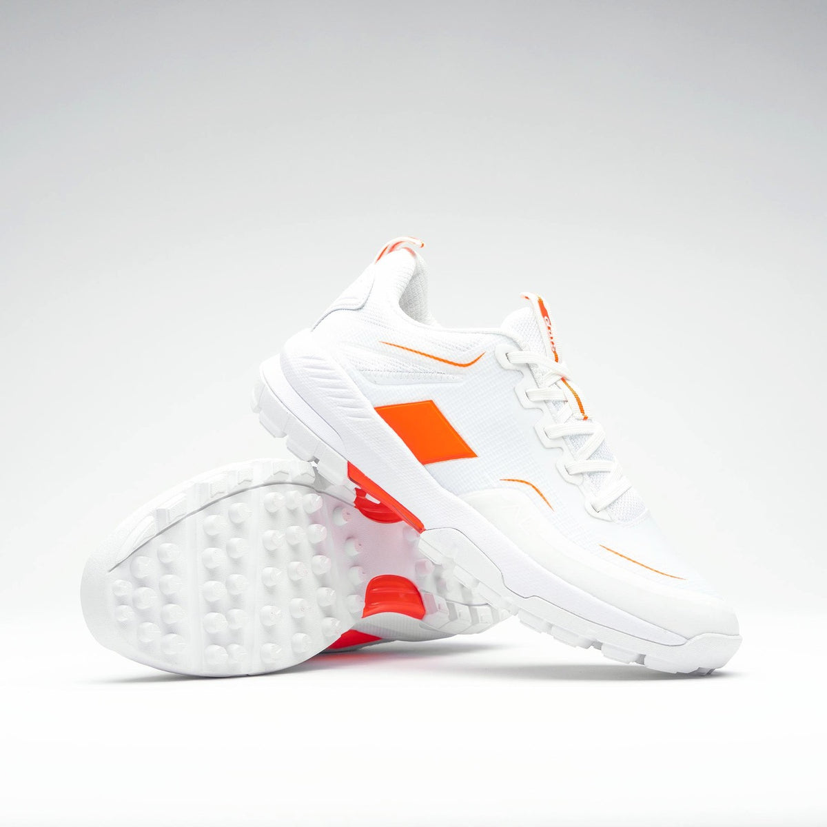 The Grays Aero Cage X Hockey Shoes by Grays are white athletic shoes with bright orange accents and a genius foam midsole for lightweight comfort. Displayed on a plain background, one shoe is upright and the other lies on its side.