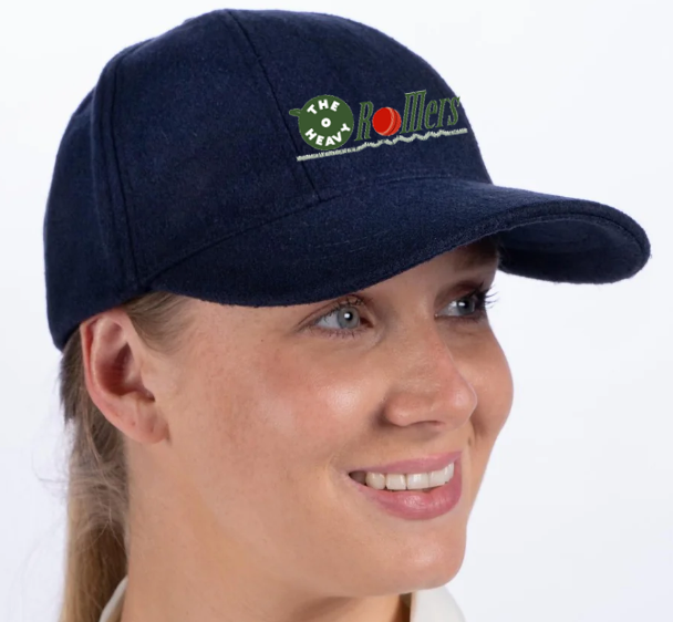 A person wearing a Gray Nicolls Heavy Rollers CC Melton County Cap in navy, senior size, with "The New Rollers" embroidered in green and red. They're smiling and gazing slightly to the side.