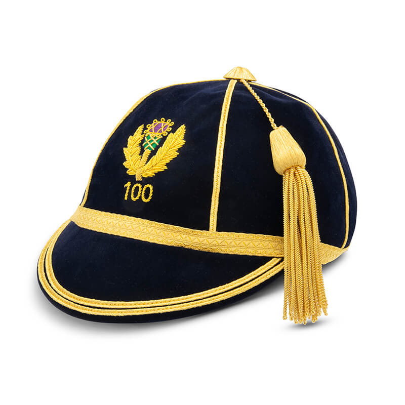 The ‘Custom, Honour & Commemorative Cap’ by Martin Berrill Sports is a navy blue velvet cap with gold trim, tassel, and an embroidered badge of leaves, a crown, and the number 100 on the front.