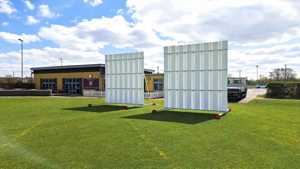 Two large white Stuart Canvas Polypropylene Sightscreens on wheels stand on a green field, with a building and parked vehicles in the background under a partly cloudy sky.