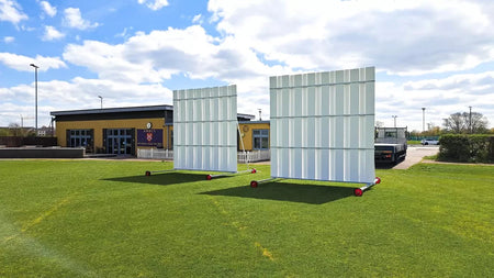 Two large white Stuart Canvas Polypropylene Sightscreens on wheels stand on a green field, with a building and parked vehicles in the background under a partly cloudy sky.