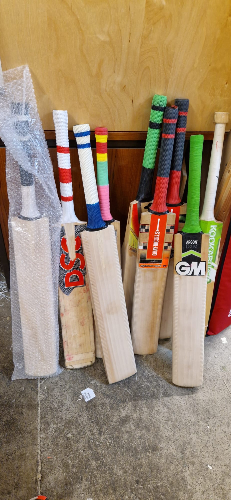 Several cricket bats with vibrant, textured handles and visible brands are lined up on a concrete floor, ready for Martin Berrill Sports’ Cricket Bat Repair - Full Refurbishment. One bat is protected in bubble wrap.