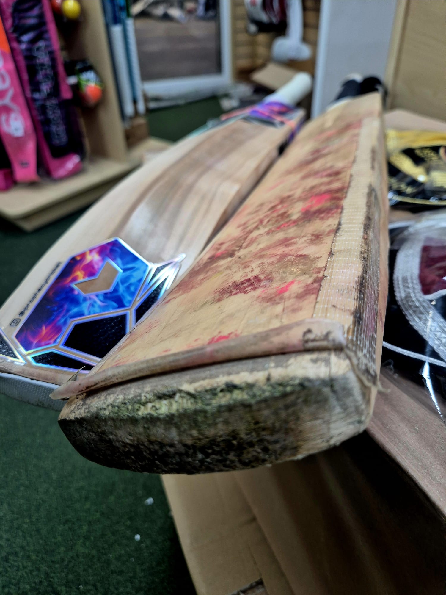 Close-up of two cricket bats on a green surface: one new with colorful stickers, the other old with visible blade damage and edge tape, featuring the Martin Berrill Sports Cricket Bat Repair - Replacement Protective Face Sheet.