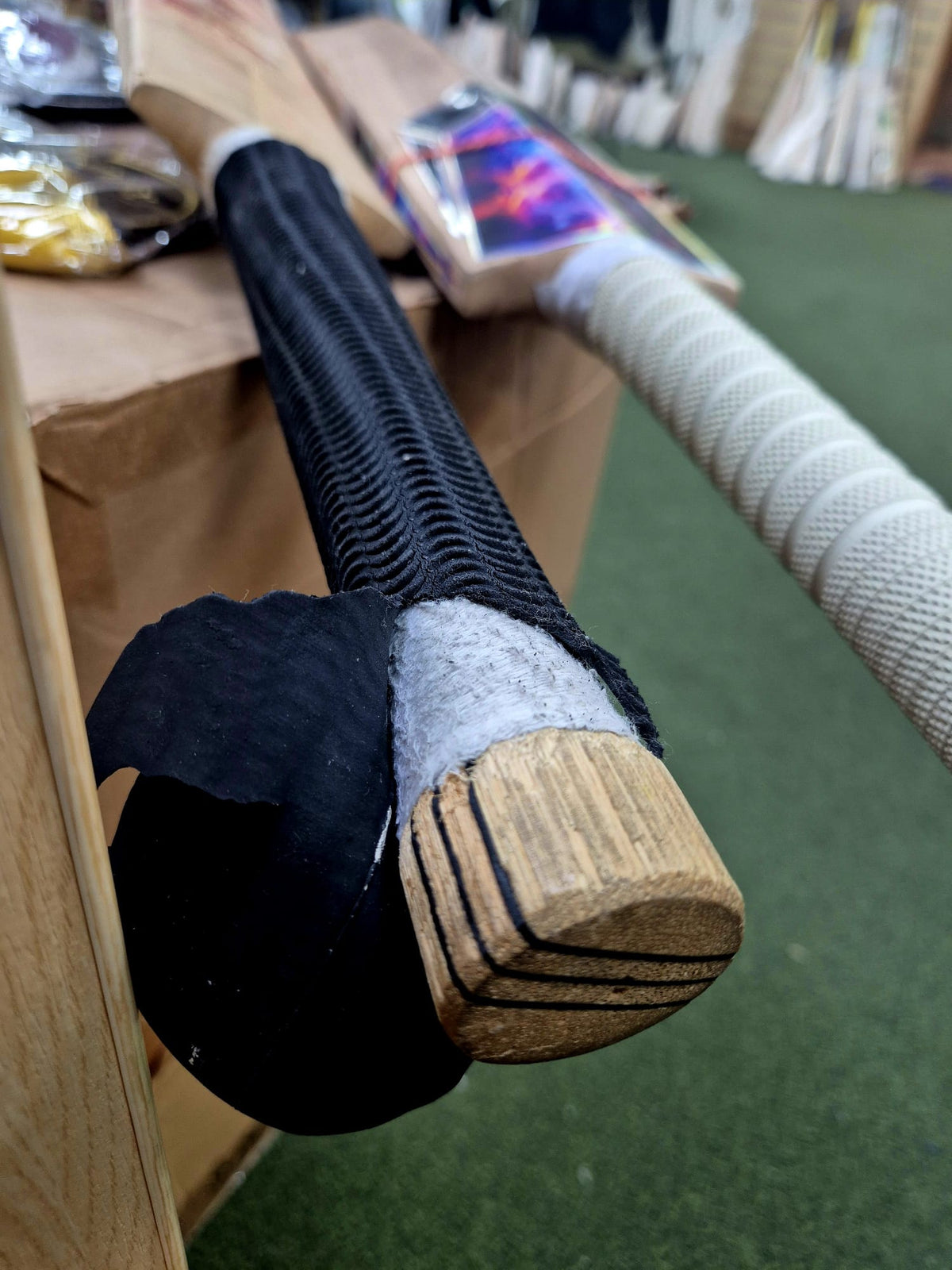 Close-up of two cricket bats with different Martin Berrill Sports Cricket Bat Replacement Grips—one black, one white—resting against a box on green turf, showing the importance of proper grip fitting for optimal performance.