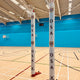 Two Gilbert Custom Printed Netball Post Protectors (Pair) with OSA and a crest stand on a light wooden court in a bright indoor gymnasium with blue walls, overhead lighting, and basketball hoops in the background.