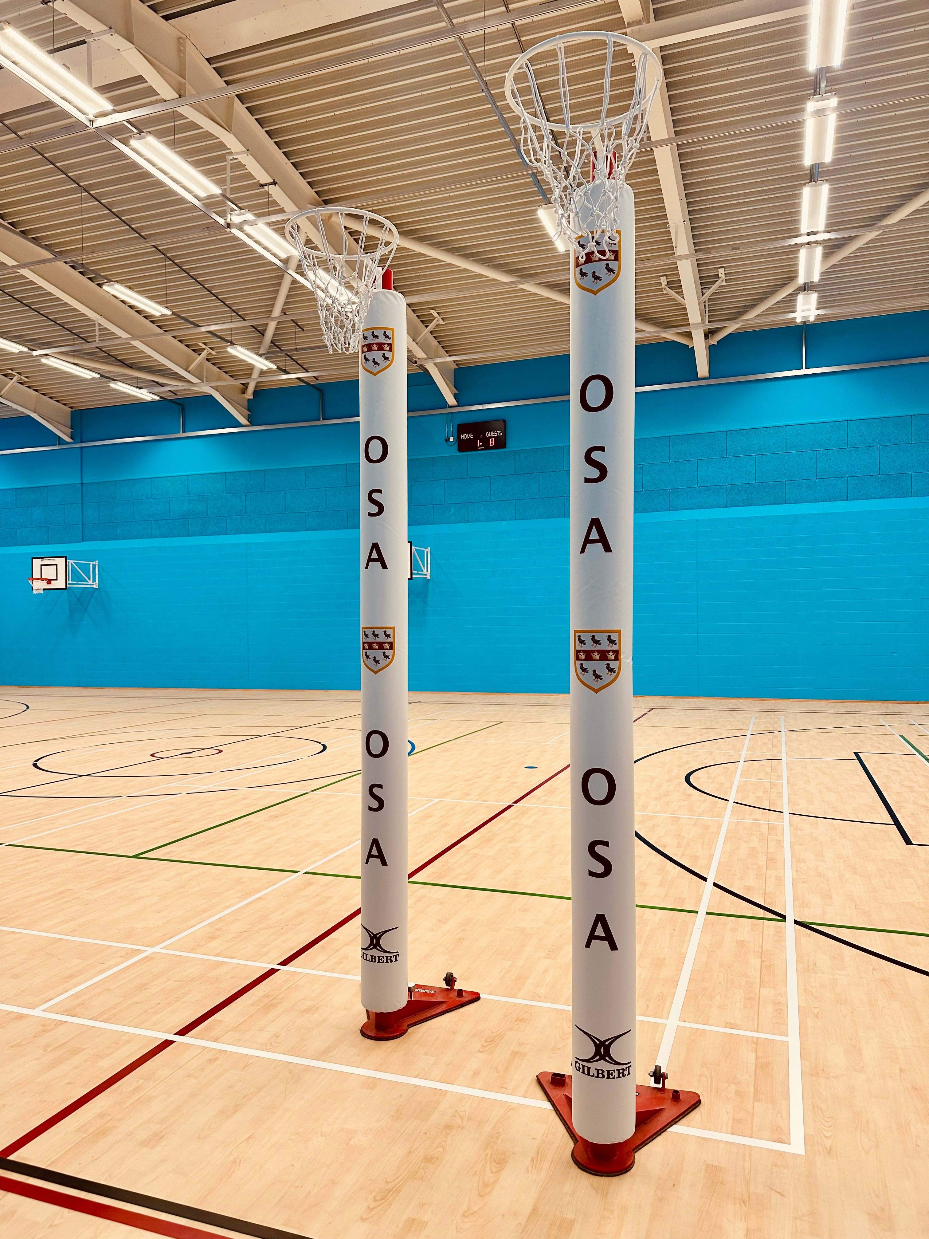 Two Gilbert Custom Printed Netball Post Protectors (Pair) with OSA and a crest stand on a light wooden court in a bright indoor gymnasium with blue walls, overhead lighting, and basketball hoops in the background.