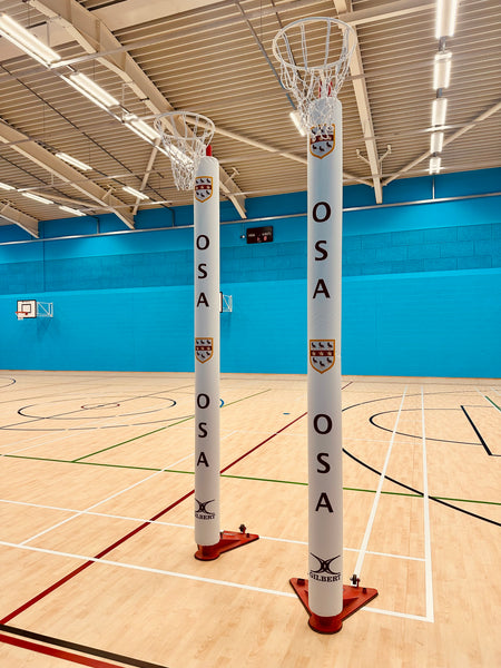 Two Gilbert Custom Printed Netball Post Protectors (Pair) with OSA and a crest stand on a light wooden court in a bright indoor gymnasium with blue walls, overhead lighting, and basketball hoops in the background.