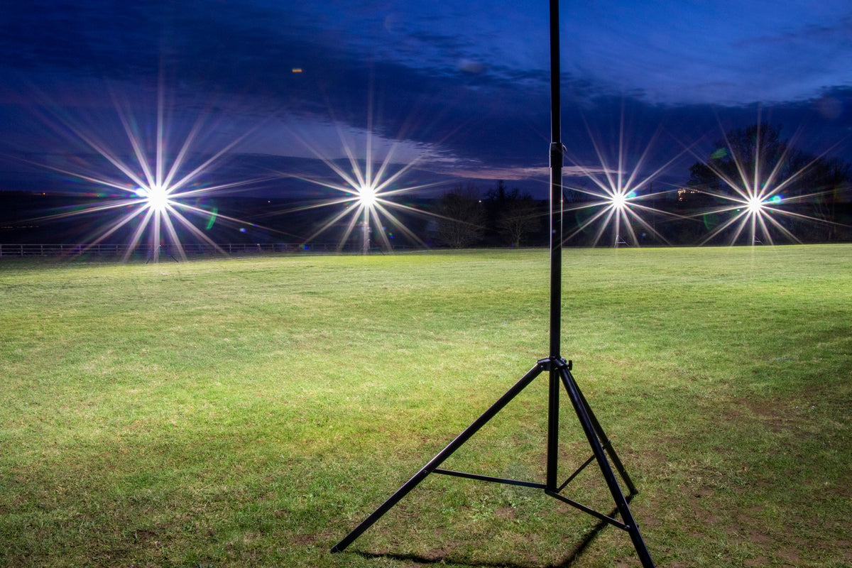 A Ritelite Sports Lite Portable Floodlights setup stands on a grassy field at dusk, with five battery-powered lights creating starburst effects in the darkening sky.