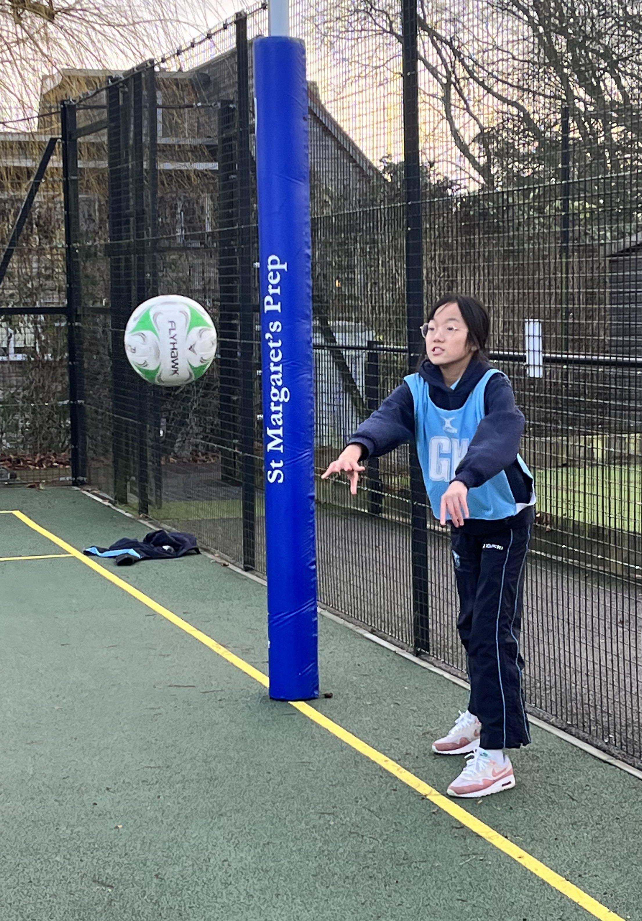 A young girl in sportswear throws a netball on an outdoor court near Gilbert Custom Printed Netball Post Protectors, branded by Gilbert and labeled St Margarets Prep, with a fence and trees visible in the background.