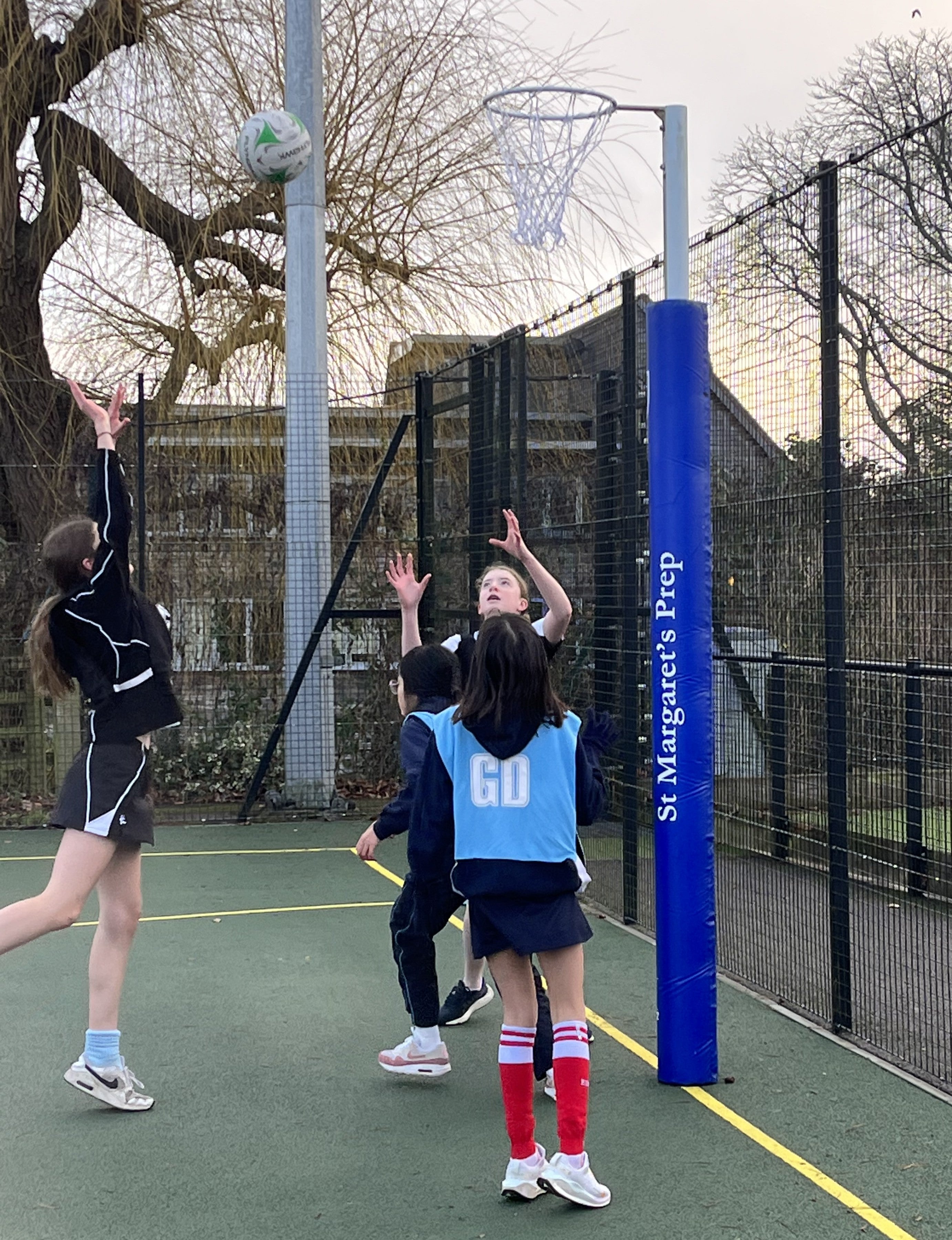 Four children play netball outdoors. One shoots as others watch near a goalpost fitted with Gilbert Custom Printed Netball Post Protectors (Pair), displaying "St. Margaret’s Prep," with trees and a building in the background.
