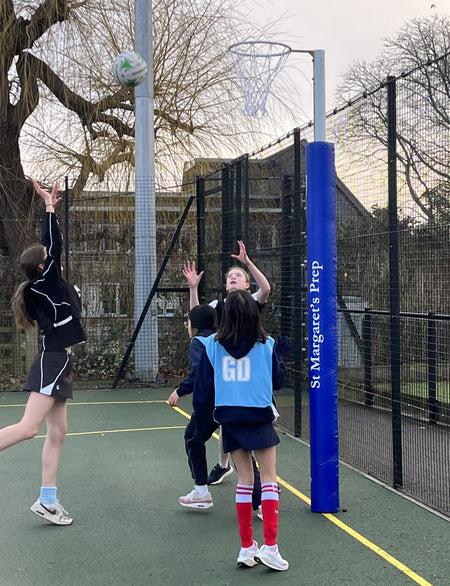 Four children play netball outdoors. One shoots as others watch near a goalpost fitted with Gilbert Custom Printed Netball Post Protectors (Pair), displaying "St. Margaret’s Prep," with trees and a building in the background.