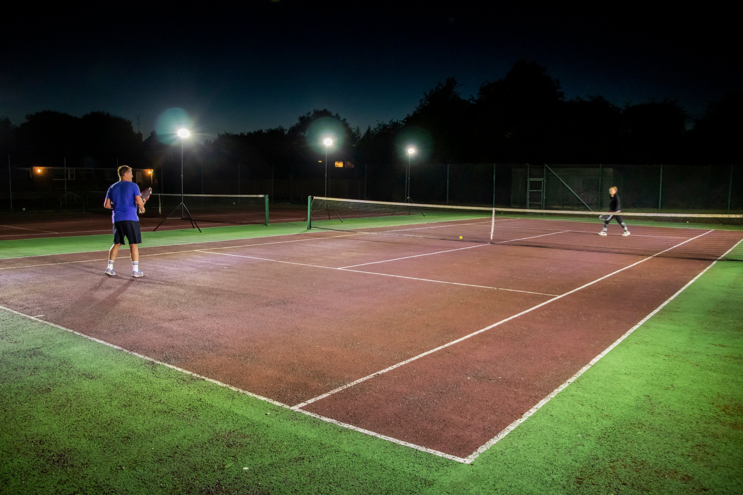 Two people play night tennis on an outdoor court, lit by bright Ritelite Sports Lite Portable Floodlights. Trees and buildings can be seen in the dark background.