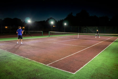 Two people play night tennis on an outdoor court, lit by bright Ritelite Sports Lite Portable Floodlights. Trees and buildings can be seen in the dark background.