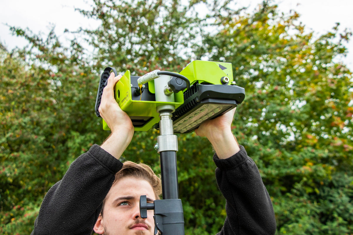 A person adjusts a green surveying instrument on a tripod outdoors, as Ritelite Sports Lite Portable Floodlights illuminate the area with leafy trees in the background.