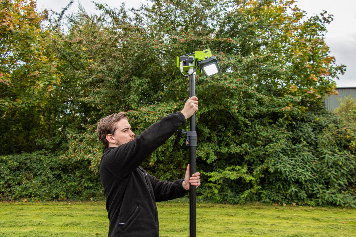 A man adjusts a Ritelite Sports Lite Portable Floodlight, powered by battery and mounted on a tall black pole, in an outdoor grassy area with dense trees and bushes in the background.
