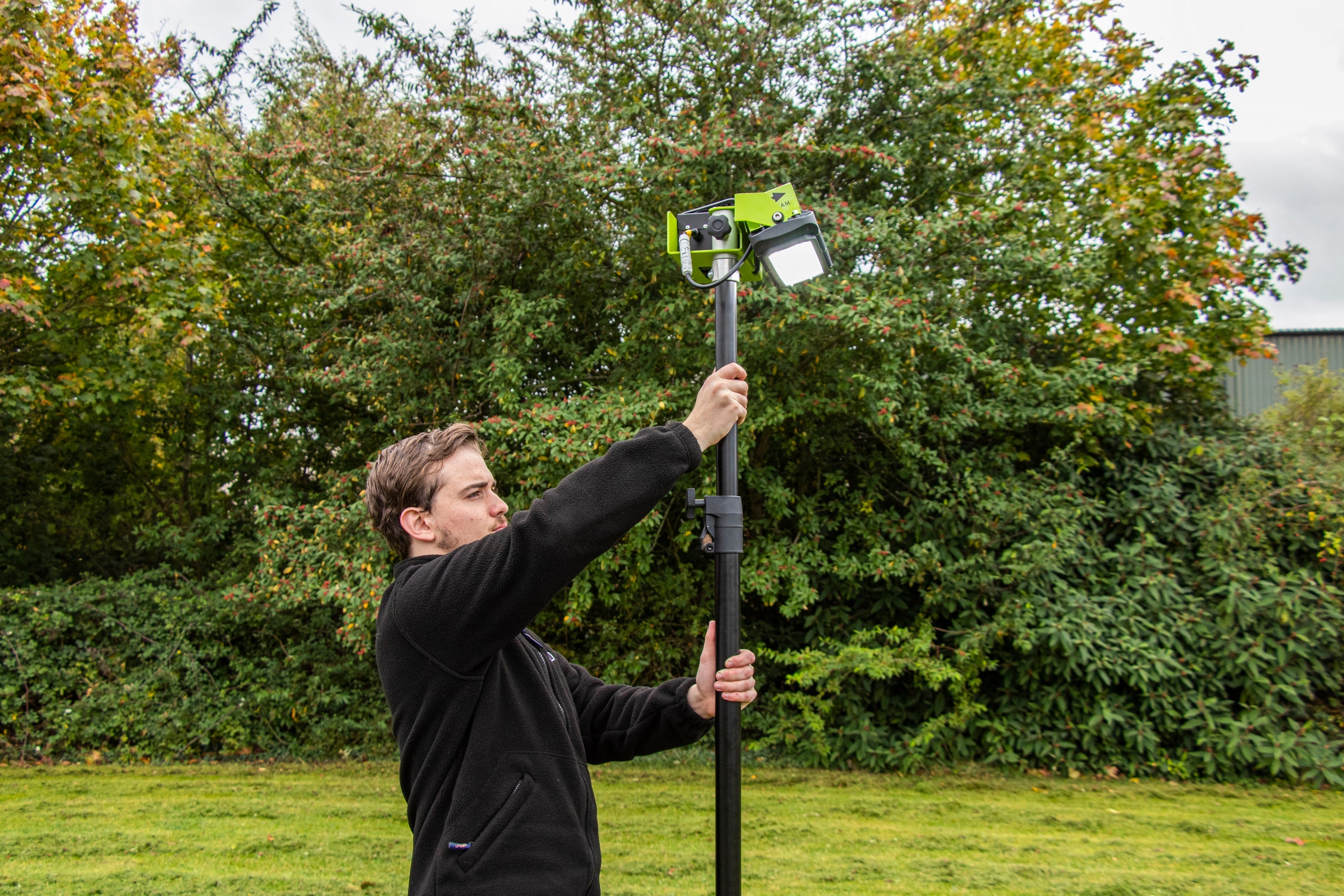 A man adjusts a Ritelite Sports Lite Portable Floodlight, powered by battery and mounted on a tall black pole, in an outdoor grassy area with dense trees and bushes in the background.