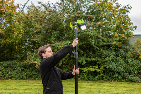 A man adjusts a Ritelite Sports Lite Portable Floodlight, powered by battery and mounted on a tall black pole, in an outdoor grassy area with dense trees and bushes in the background.