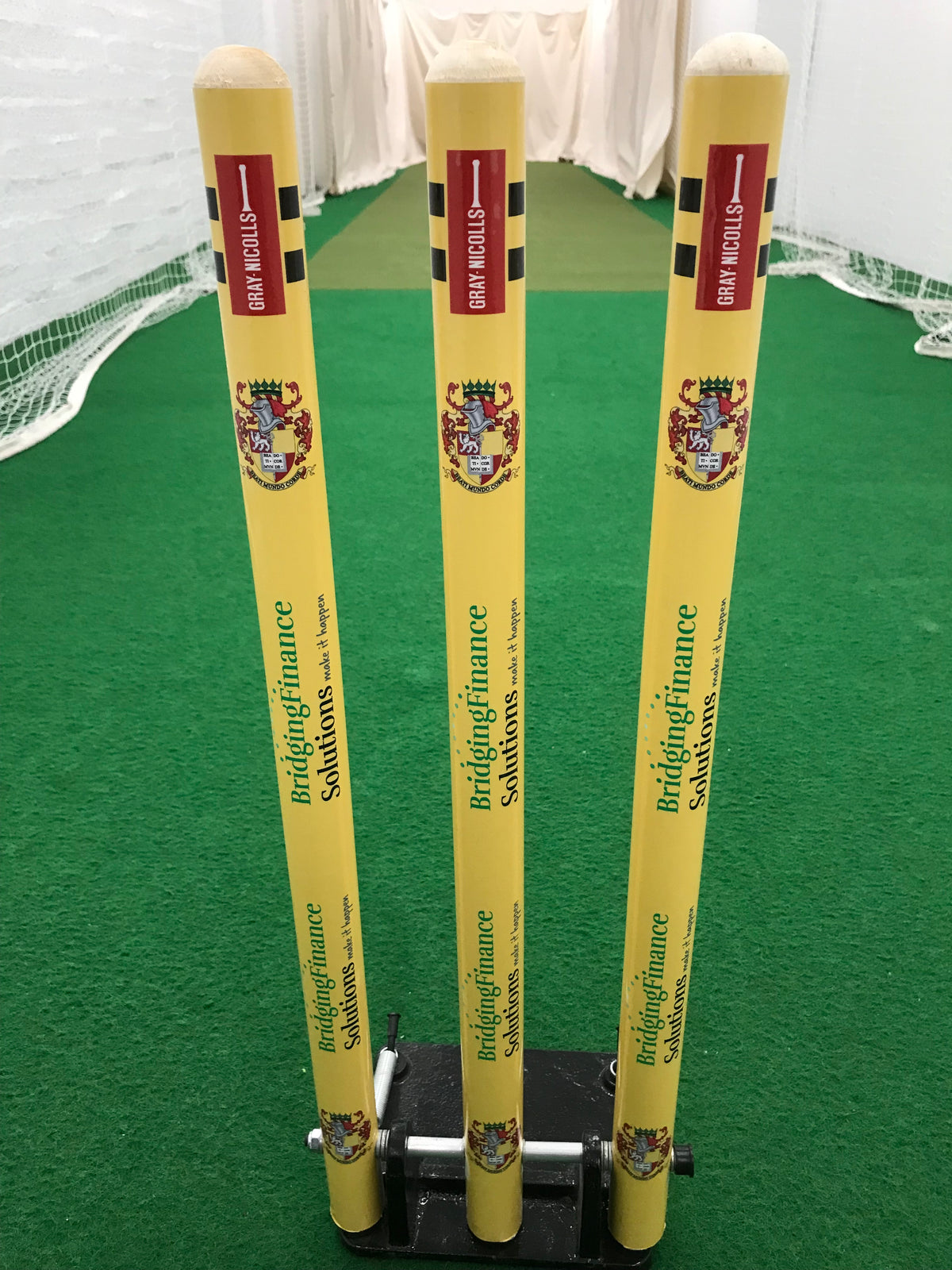 Three bright yellow Custom Printed Spring Back Stumps by Gray Nicolls stand on indoor green turf with white netting and walls behind. They're ideal cricket training stumps for practice sessions.
