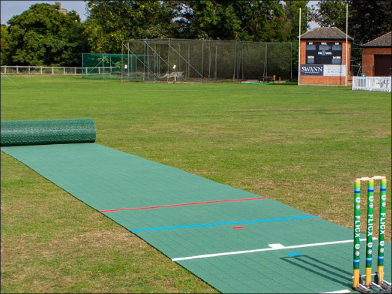 The Flicx UK Flicx 2G MAP (Multi Age Pitch) Match Cricket Pitch is rolled out on a field with stumps, set near a scoreboard and netted area—ideal for clubs and schools.